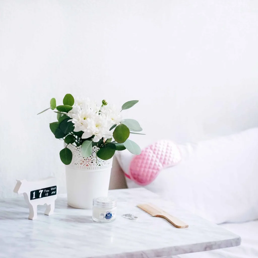 bed with sleep mask, side table with pot of flowers, cream, comb