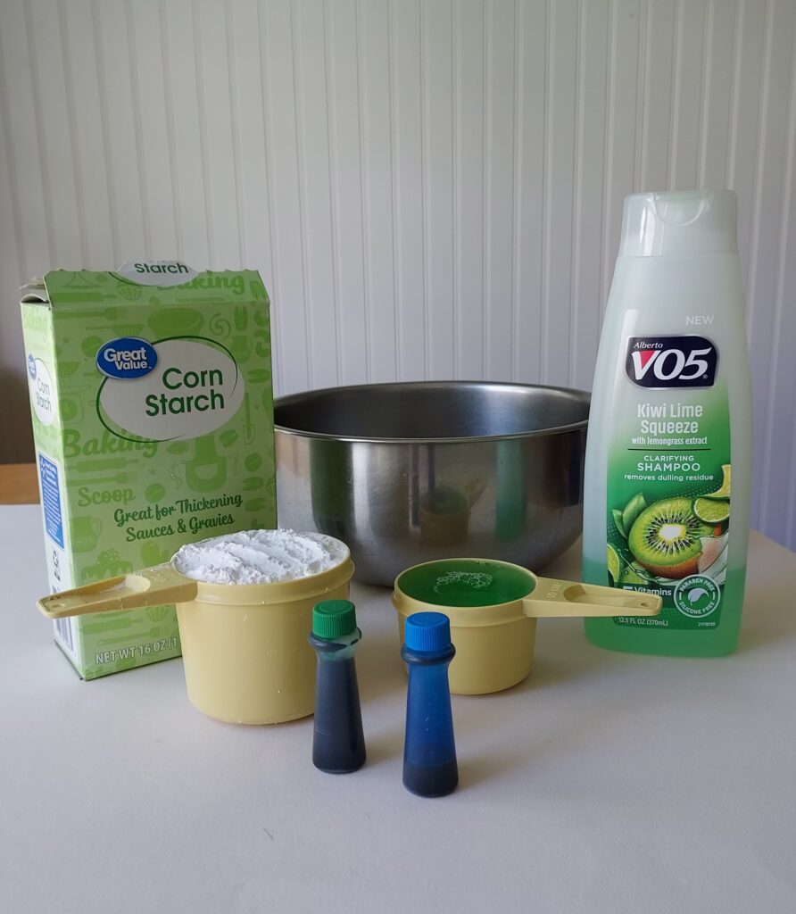 photo of a box of cornstarch, metal mixing bowl, bottle of shampoo, 1 tube of green food coloring, 1 tube of blue food coloring, 1 cup measuring cup filled with cornstarch, 1/2 cup measuring cup filled with green shampoo, all to make slime without using glue