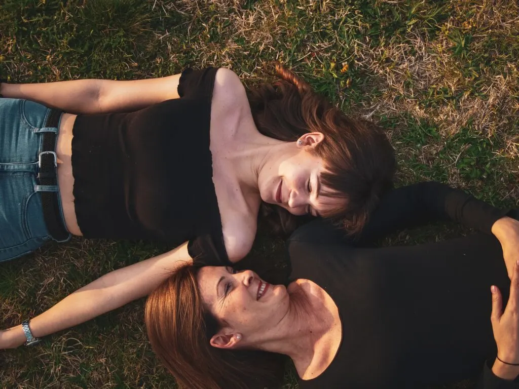 Two women laying in the grass facing each other smiling.  They appear to be a mother and young adult daughter.