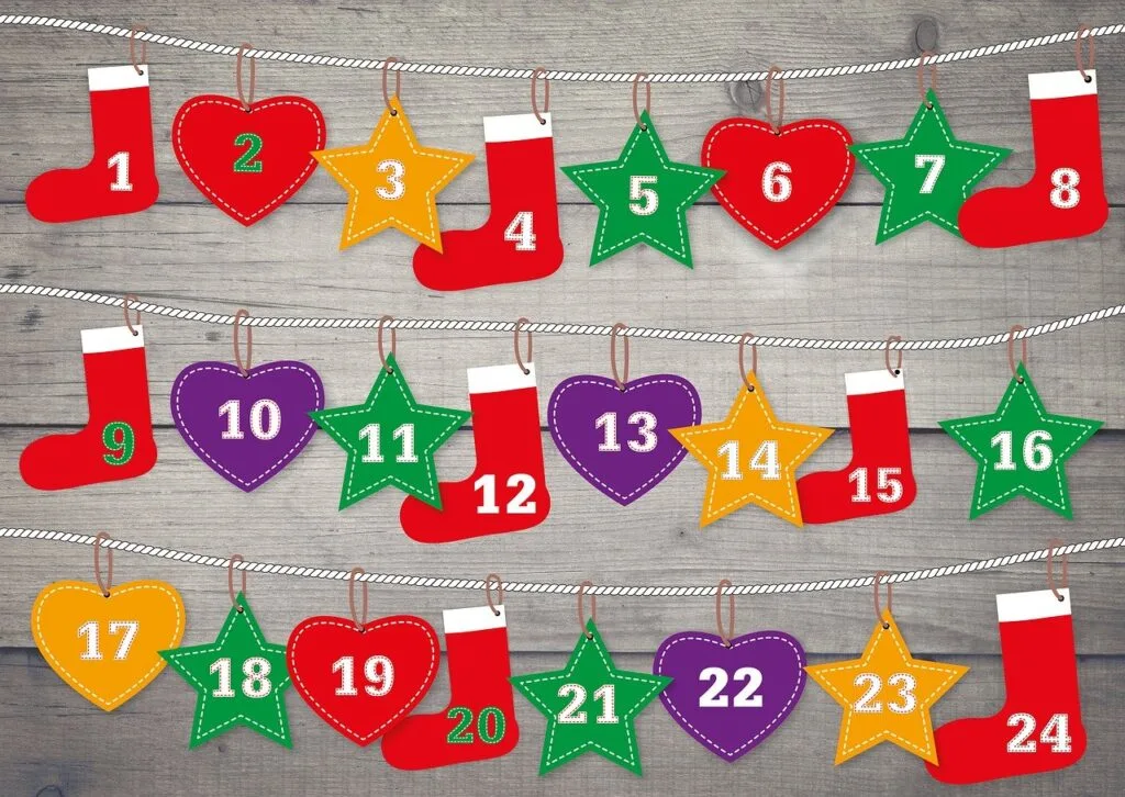 gray wooden board background three rows of white cord. Red, green, yellow and purple stars, hearts, and stockings, each labeled with numbers 1-24 for advent calendar