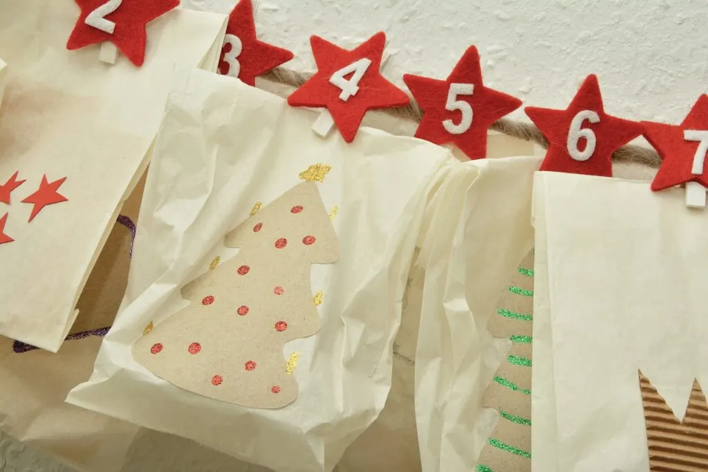 White paper bags decorated for Christmas attached to rope by clothespins. Red stars with numbers are covering the clothes pins. Idea for advent calendar