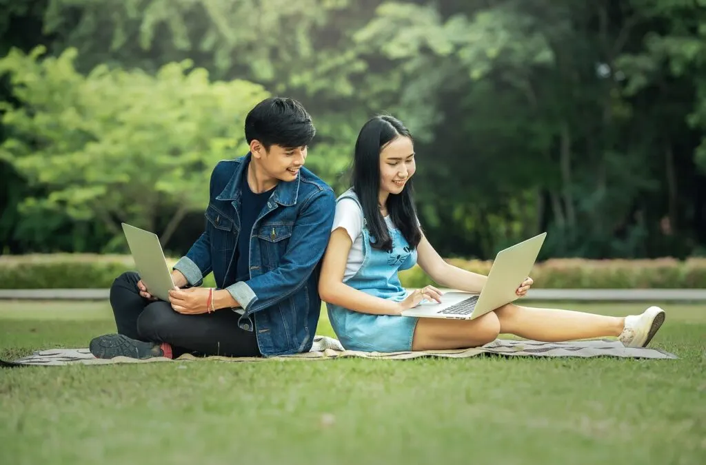 date ideas for teenagers, two teens sitting on a blanket in the grass back to back holding laptops